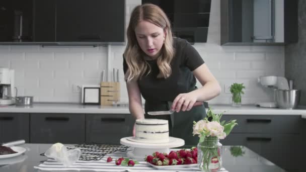 Professional chef is cooking cake. Young attractive housewife uses metal spatula and rotating table to aligns white cream on chocolate cake — Stock Video