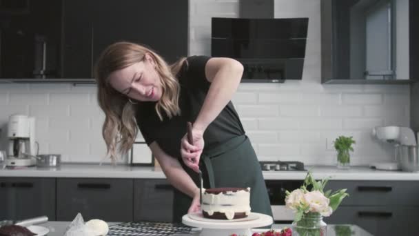 Profi-Koch kocht Kuchen. Junge attraktive Hausfrau richtet weiße Sahne mit Metallspachtel und Drehtisch auf Schokoladenkuchen aus — Stockvideo