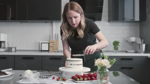 Profi-Koch kocht Kuchen. Junge attraktive Hausfrau richtet weiße Sahne mit Metallspachtel und Drehtisch auf Schokoladenkuchen aus — Stockvideo