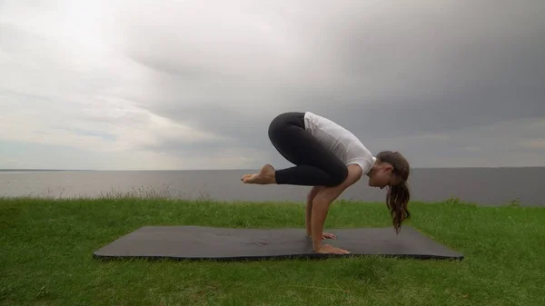 Jovem mulher apto praticar ioga na costa perto do lago ou do mar. Mulher fazendo Crane Bakasana pose — Fotografia de Stock