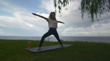 Genç, formda bir kadın göl ya da deniz kıyısında yoga yapıyor. Savaşçı 2 pozu veren kadın.