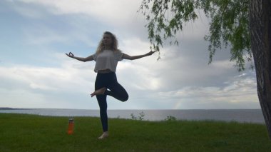 Genç ve formda bir kadının göl ya da denizin kıyısında yoga yapışının arkası. Kadın ayakta yarım lotus pozu veriyor.