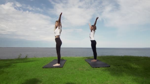 İki genç kadın göl ya da deniz kıyısında yoga yapıyor. Dans eğitmeni pozu.. — Stok video