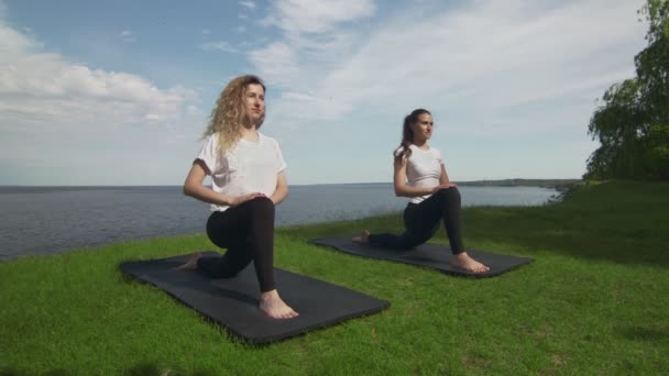 Due giovani donne praticano yoga sulla costa vicino al lago o al mare. Formazione Crescent Lunge sulla posa del ginocchio . — Video Stock