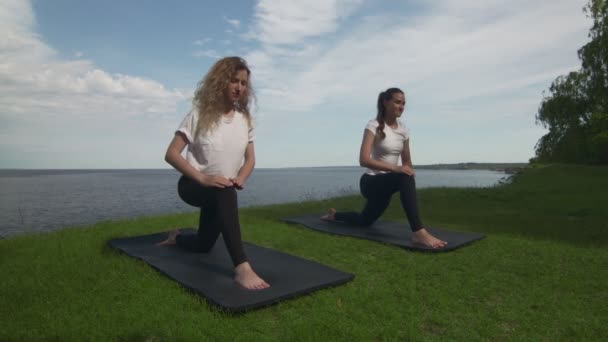 Två unga kvinnor utövar yoga på kusten nära sjön eller havet. Träning Crescent Lunge på knät pose. — Stockvideo