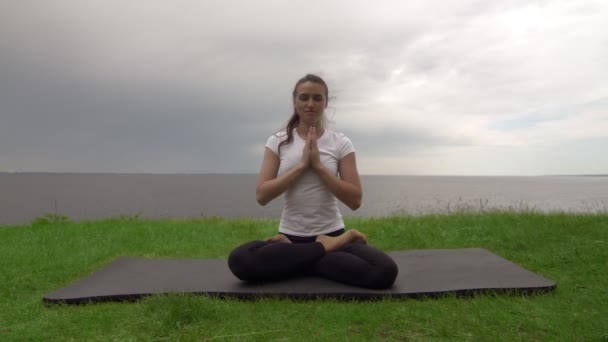 Young fit woman practice yoga on coast near the lake or sea. Woman sitting in lotus pose and meditating — Stock Video