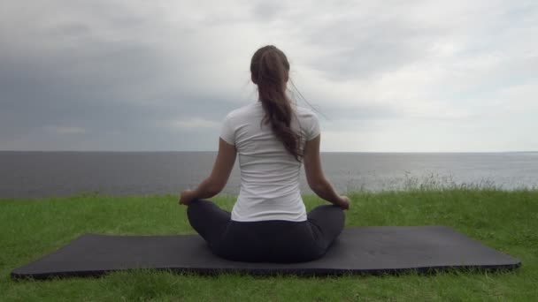 Young fit woman practice yoga on coast near the lake or sea. Woman sitting in lotus pose and meditating — Stock Video