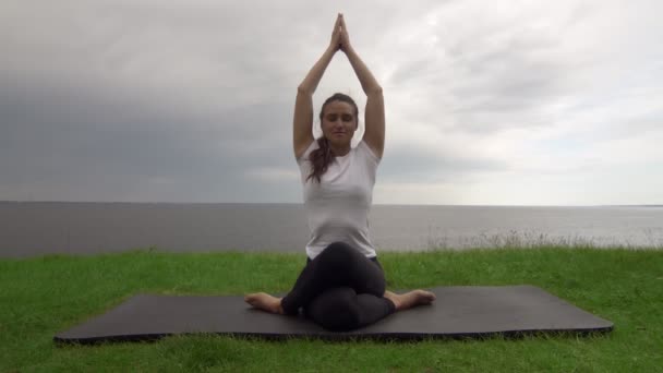 Mladá fit žena cvičí jógu na pobřeží u jezera nebo u moře. Žena sedící v kolenou Pózuje a medituje — Stock video