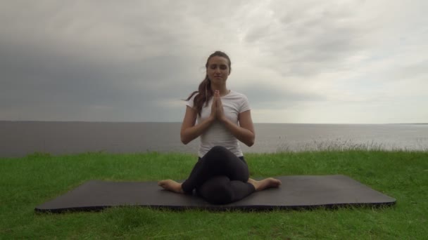 Mladá fit žena cvičí jógu na pobřeží u jezera nebo u moře. Žena sedící v kolenou Pózuje a medituje — Stock video