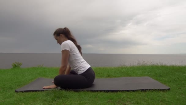 Mladá fit žena cvičí jógu na pobřeží u jezera nebo u moře. Žena sedí v přední splits hanumanasana póza — Stock video