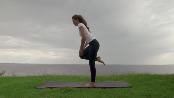 Ung vältränad kvinna utövar yoga på kusten nära sjön eller havet. Kvinna gör balansering stående splittringar och sedan stående framåt böja Poser — Stockvideo