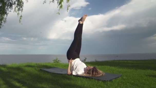 Junge, fitte Frauen praktizieren Yoga an der Küste in der Nähe des Sees oder Meeres. Frau macht unterstützte Schulterstandhaltung — Stockvideo