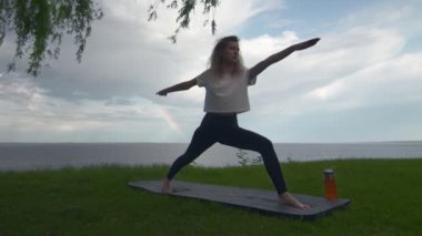 Genç, formda bir kadın göl ya da deniz kıyısında yoga yapıyor. Kadın birinci ve ikinci pozisyonları yapıyor.