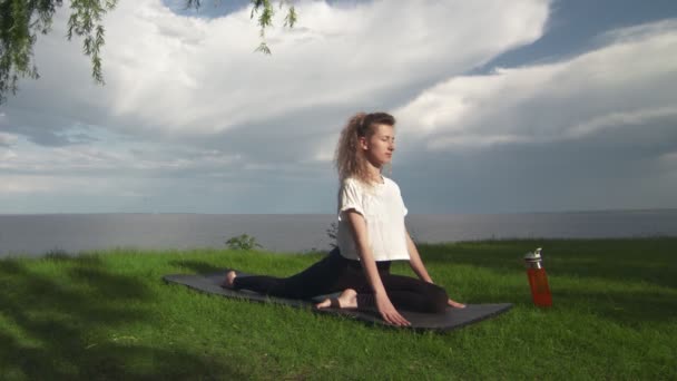 Fiatal nő gyakorolja jóga a parton, közel a tó vagy a tenger. Nő csinál Half Pigeon Pose — Stock videók