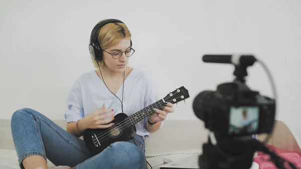 Young attractive girl blogger with headphones on head is recording video with camera, singing and playing on guitar — Stock Photo, Image