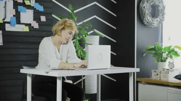 Young woman is talking on her smartphone while typing on notebook — Stock Photo, Image