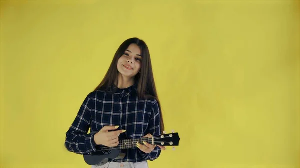 Menina jovem cantando e tocando ukulele no fundo amarelo no estúdio — Fotografia de Stock