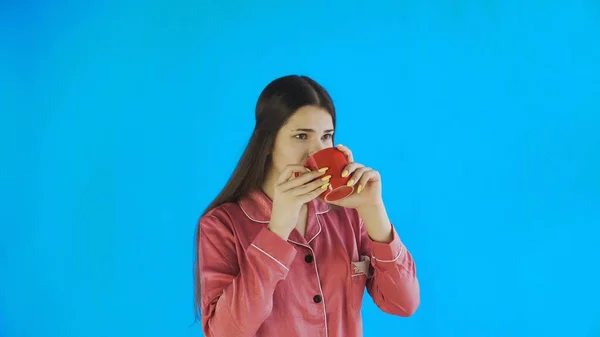 Mujer joven en pijama está tomando té o café sobre fondo azul en el estudio —  Fotos de Stock