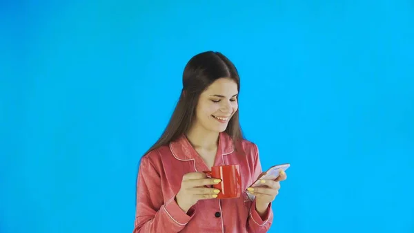 Chica feliz en pijama es beber té o café y el uso de teléfono sobre fondo azul en el estudio —  Fotos de Stock
