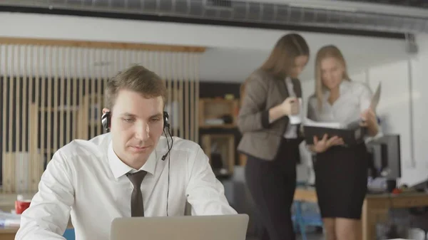 Man is consulting clients by phone, sitting in an office in daytime, two women on blur background — Stock Photo, Image