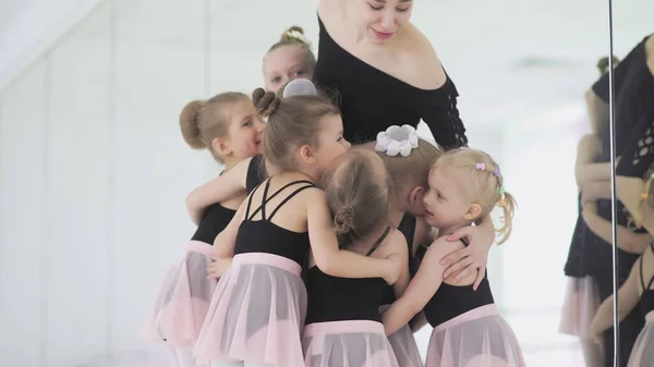 Young ballet teacher sitting on bench in ballet school with little girls in black leotards and communicating with them