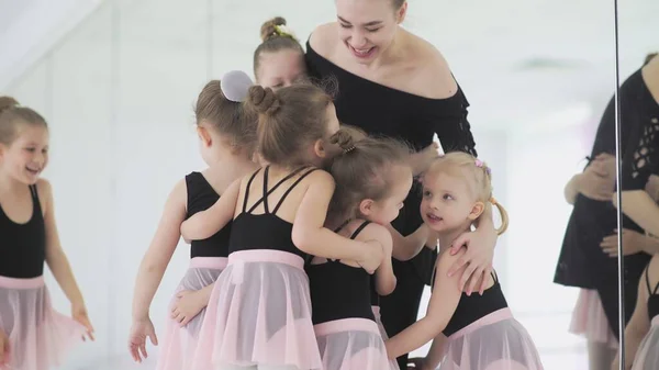 Young ballet teacher sitting on bench in ballet school with little girls in black leotards and communicating with them