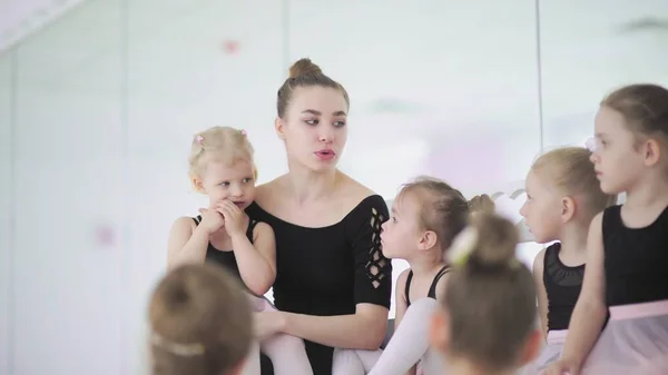 Young ballet teacher sitting on bench in ballet school with little girls in black leotards and communicating with them