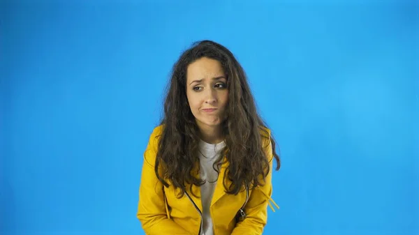 Triste réfléchie jeune belle femme en veste jaune debout dans Studio avec fond bleu. — Photo