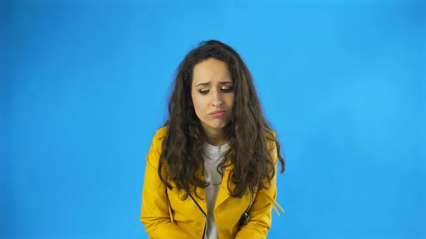 Triste pensativo jovem mulher bonita em casaco amarelo de pé no estúdio com fundo azul . — Fotografia de Stock