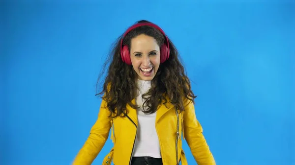 노란 자켓을 입고 춤추는 젊은 여자 가 스튜디오에서 파란색 배경으로 헤드폰으로 음악을 듣고 있는 모습. — 스톡 사진
