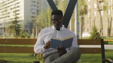 Gözlüklü genç Afrikalı öğrenci parkta oturuyor, kitap okuyor ve kahve içiyor.