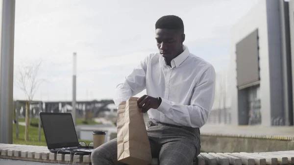 Joven hombre de negocios afroamericano relajándose y comiendo croissant en el banco en el parque de la ciudad — Foto de Stock