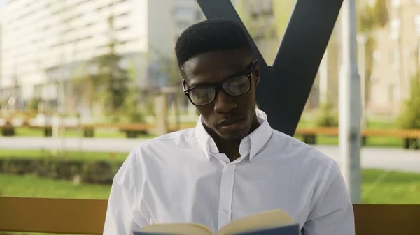 Jovem estudante africano de óculos está sentado no parque, lendo livro e bebendo café — Fotografia de Stock