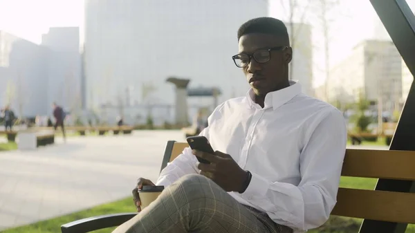 若いアフリカ系アメリカ人の男は、オフィスの近くの公園でスマートフォンやコーヒーや紅茶でアプリを使用して — ストック写真