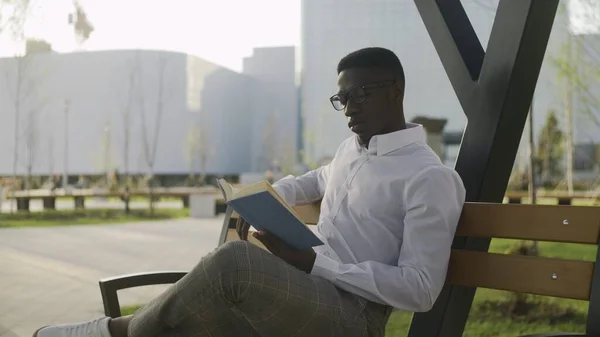 Gözlüklü genç Afrikalı öğrenci parkta oturuyor, kitap okuyor ve kahve içiyor.