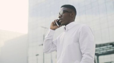 Gözlüklü Afrikalı Amerikalı İşadamı cep telefonuyla konuşuyor, ofisinin yanında duruyor.