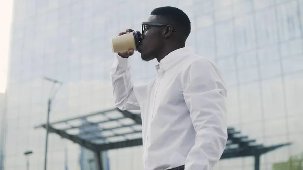 Стильный афроамериканский бизнесмен в белой рубашке пьет кофе на улице возле офиса — стоковое фото
