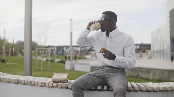 Jovem afro-americano bebendo café ou chá e comendo sanduíche no parque perto do escritório — Fotografia de Stock