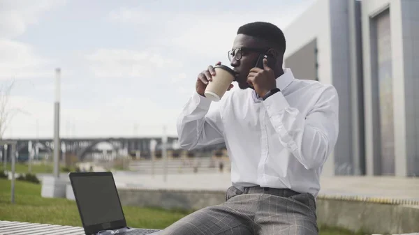 Atractivo hombre afroamericano usando smartphone y tomando café o té en el parque cerca de la oficina — Foto de Stock