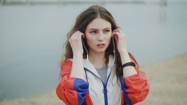 Mujer joven está usando auriculares para la motivación musical para el entrenamiento cardiovascular, río o mar en el fondo —  Fotos de Stock