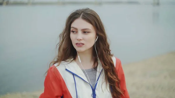 Mujer joven está usando auriculares para la motivación musical para el entrenamiento cardiovascular, río o mar en el fondo —  Fotos de Stock