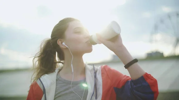Esmer kız parkta koştuktan sonra soğuk su içiyor. Spor yaptıktan sonra su içen güzel sporcu kadın sağlık ve spor, parlayan ışık.