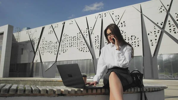 Woman is calling by phone and looking on display of laptop thoughtfully outdoor — Stock Photo, Image