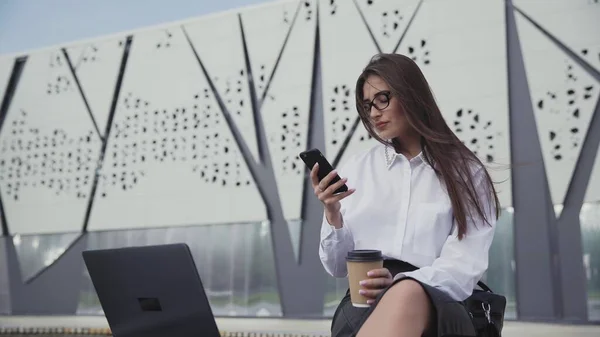 Beautiful business woman in glasses drinking coffee and looking at her smartphone — Stock Photo, Image