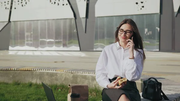 Красива ділова жінка їсть круасан під час розмови на мобільному телефоні і посміхається на відкритому повітрі — стокове фото