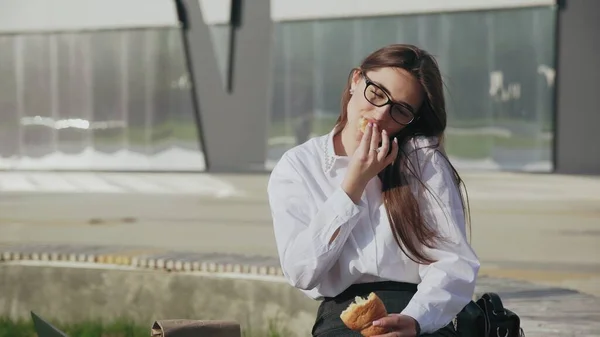 A successful corporate woman eating a croissant while talking on the mobile phone and smiling outdoors — Stock Photo, Image