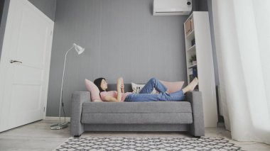 Beyaz tenli genç bir kadın evdeki kanepede uzanırken akıllı telefonuyla tıkırdıyor, kaydırıyor ve daktilo kullanıyor.
