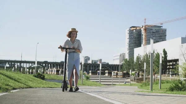 Atractiva mujer en sombrero de paja caminando con ella la patada eléctrica scooter — Foto de Stock