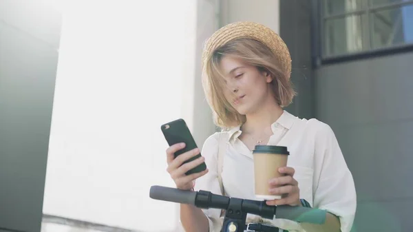 Woman is using smartphone and drinking coffee after ride on electric kick scooter — Stock Photo, Image