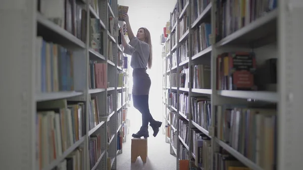 Jovem estudante menina tomando livro de prateleira na biblioteca da universidade . — Fotografia de Stock